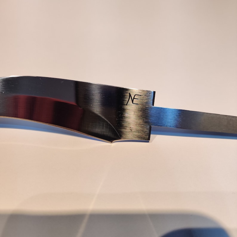K&ouml;nnte beinhalten: Nahaufnahme einer silbernen Messerklinge mit einer gebogenen Kante. Die Klinge hat eine geb&uuml;rstete Oberfl&auml;che und die Buchstaben "NE" sind in das Metall einge&auml;tzt.