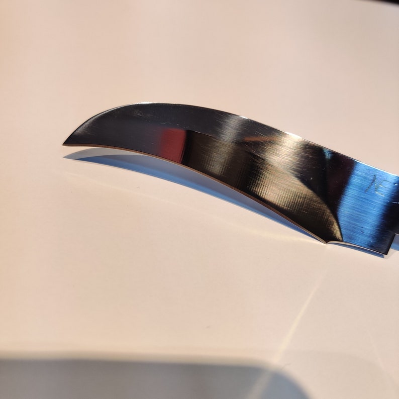 K&ouml;nnte beinhalten: Nahaufnahme einer gl&auml;nzenden, silbernen Messerklinge mit einer gebogenen Kante. Die Klinge reflektiert das Licht und hat eine dezente, geb&uuml;rstete Oberfl&auml;che.