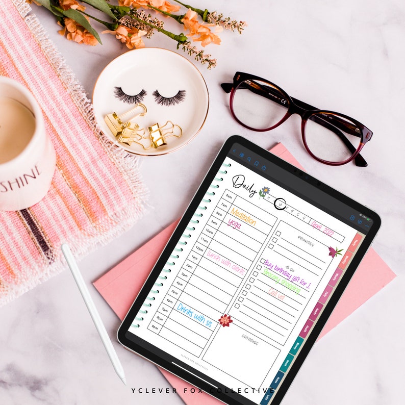 May include: An iPad showing a digital daily planner with a floral design. The planner includes a schedule with times and tasks like "Meditation" and "Drinks with sis." Other items include a white stylus, glasses, and a decorative dish with eyelashes.