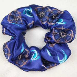 Op de afbeelding: Een donkerblauwe scrunchie met een herhalend bloemmotief in bruin- en tinten. De stof heeft een satijnglans en de scrunchie is geplooid voor een volumineuze look. Een modieus haaraccessoire.