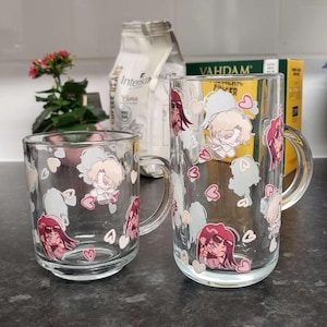 May include: Two clear glass mugs with handles, both decorated with a white background and pink hearts. The mugs feature cartoon illustrations of people with pink and white accents. The mugs are on a black countertop.