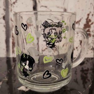 Puede incluir: Taza de vidrio transparente decorada con personajes de estilo anime y formas de corazón en negro y verde. La taza tiene un asa y es adecuada para bebidas frías o calientes. El diseño incluye notas musicales.