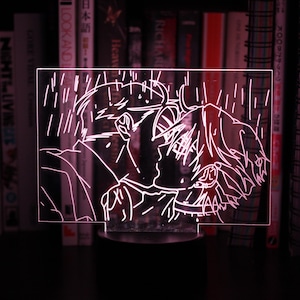 Puede incluir: Una luz nocturna acrílica brillante con una ilustración romántica de estilo anime de dos figuras besándose bajo la lluvia. La luz emite un suave tono rosa, con el diseño grabado en blanco. La lámpara se encuentra sobre una base negra, con libros en el fondo.