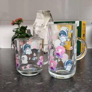 May include: Two glass mugs with handles, featuring cartoon characters and hearts. The mugs are on a dark countertop.