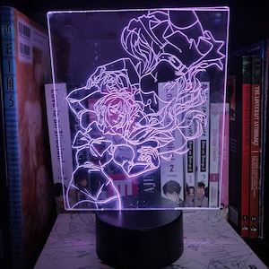 May include: A pink LED night light with a white outline of two anime characters. The characters are a woman with long hair and a man with short hair. The light is on a black base.
