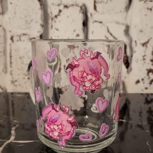 May include: Clear glass mug decorated with pink anime-style character illustrations and heart shapes. The mug features a black handle and is set against a white brick wall background.