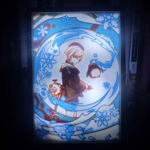 Pode incluir: Obra de arte iluminada com um personagem em estilo anime dentro de um design circular azul e branco. A obra de arte inclui motivos de flocos de neve e peixes, com texto japonês no canto inferior esquerdo. O personagem usa uma roupa marrom e bege.