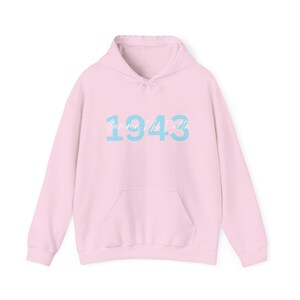 May include: A light pink hooded sweatshirt with a front pocket and a drawstring hood. The sweatshirt features the text "Gamma Phi Beta" above the number "1943" in light blue. The sweatshirt is made of a soft material.