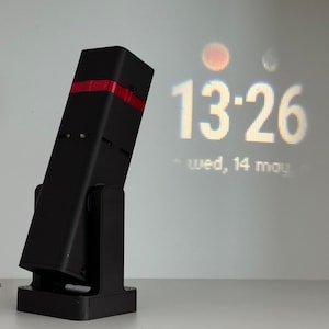 May include: A black rectangular projector with a red accent on top, mounted on a black stand. The projector is angled towards a white wall, displaying the time 13:26 and the date.