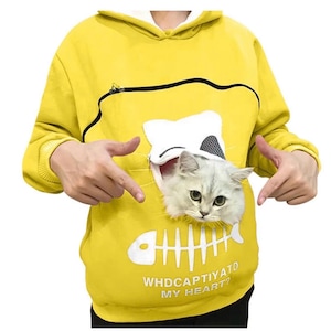 Puede incluir: Una sudadera con capucha de color amarillo brillante que presenta un bolsillo frontal grande con una abertura de cremallera en forma de cabeza de gato. Un gato blanco esponjoso asoma por el bolsillo. Debajo hay un gráfico de espina de pescado y el texto WHDCAPTIYATD MY HEART?