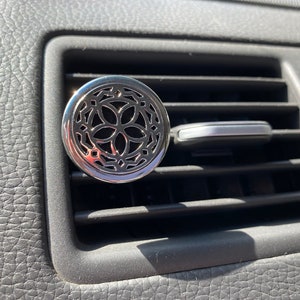 May include: A silver car air freshener with a decorative, geometric design. The air freshener is attached to a car's air vent.