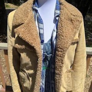 May include: A tan suede jacket with a shearling collar and lining. The jacket has two front pockets and is worn over a blue plaid shirt and denim overalls. The jacket is displayed on a mannequin.