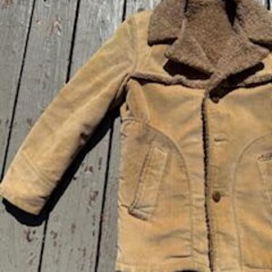 May include: A tan corduroy jacket with a brown faux-fur collar and lining. The jacket has a button-up front, two front pockets, and a classic design. The jacket is displayed on a weathered wooden surface.
