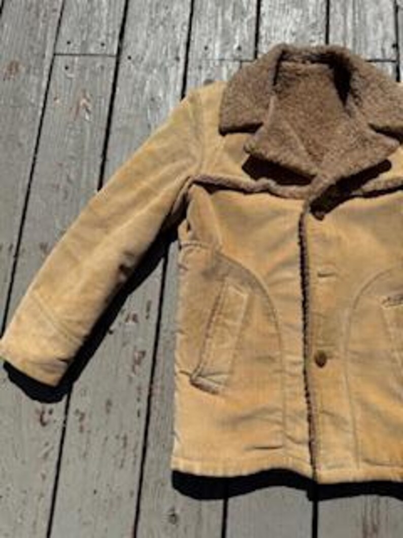 May include: A tan corduroy jacket with a brown faux-fur collar and lining. The jacket has a button-up front, two front pockets, and a classic design. The jacket is displayed on a weathered wooden surface.