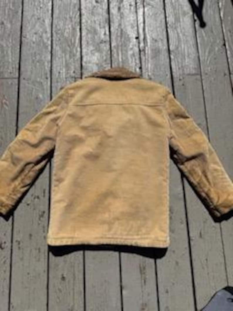 May include: Tan corduroy jacket with a brown faux fur collar. The jacket has a button-up front, two chest pockets, and long sleeves. The jacket is shown from the back, with the collar and shoulders in focus.