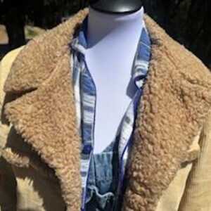 May include: A tan corduroy jacket with a brown faux fur collar and lining. The jacket is worn over a blue and white plaid shirt and denim overalls. The jacket has a classic, vintage style.