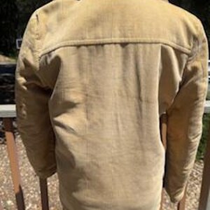 May include: Tan corduroy jacket with a shearling collar. The jacket has a button-up front and two chest pockets. The back of the jacket is visible, showing the yoke and vertical corduroy lines. The jacket is worn over blue jeans.