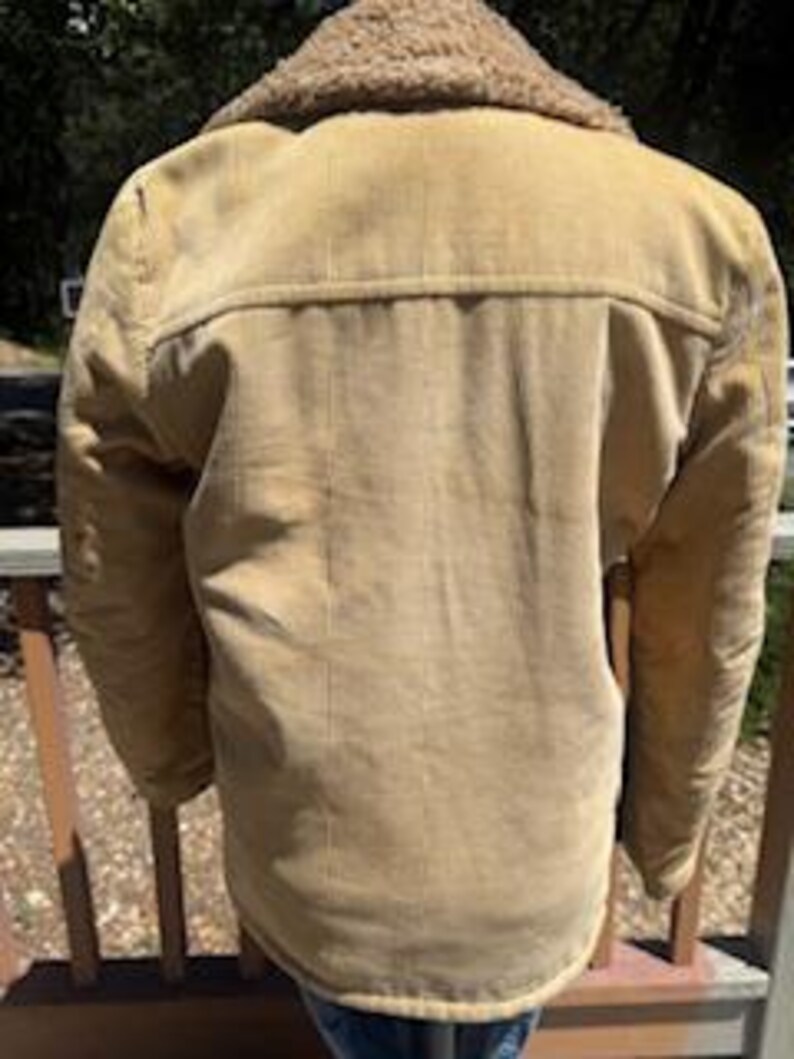 May include: Tan corduroy jacket with a shearling collar. The jacket has a button-up front and two chest pockets. The back of the jacket is visible, showing the yoke and vertical corduroy lines. The jacket is worn over blue jeans.