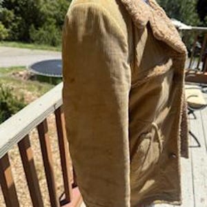 May include: A tan corduroy jacket with a shearling collar and lining. The jacket has two front pockets and a button closure. The jacket is shown on a wooden deck with a natural background.
