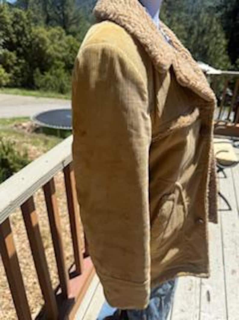 May include: A tan corduroy jacket with a shearling collar and lining. The jacket has two front pockets and a button closure. The jacket is shown on a wooden deck with a natural background.