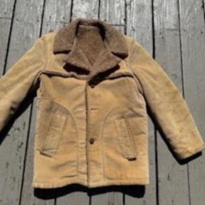 May include: A tan corduroy jacket with a brown faux fur collar and lining. The jacket has two front pockets and a button closure. The jacket is laid out on a weathered wooden surface.