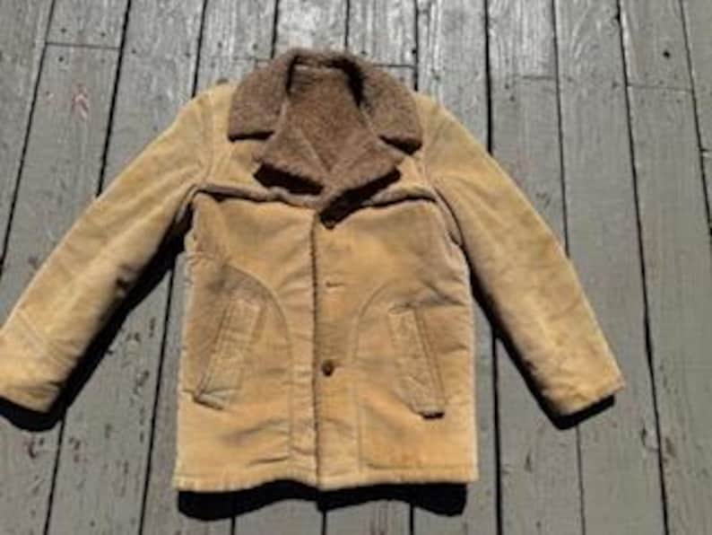 May include: A tan corduroy jacket with a brown faux fur collar and lining. The jacket has two front pockets and a button closure. The jacket is laid out on a weathered wooden surface.