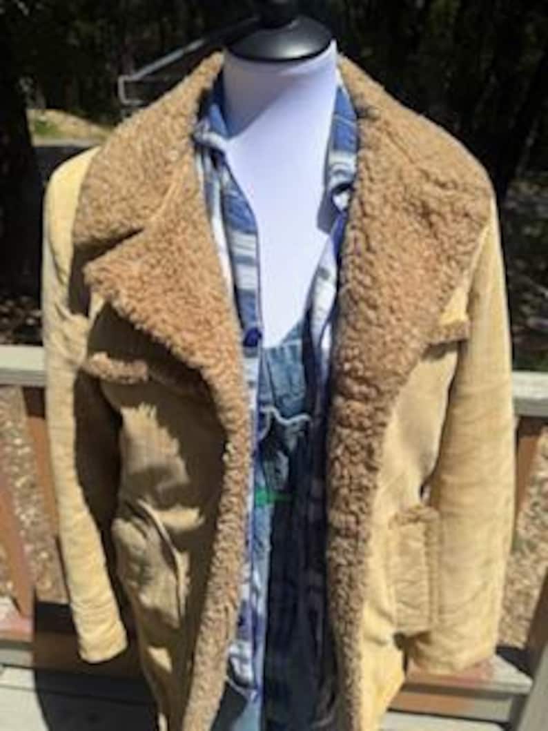May include: A tan suede jacket with a shearling collar and lining. The jacket has two front pockets and is worn over a blue plaid shirt and denim overalls. The jacket is displayed on a mannequin.