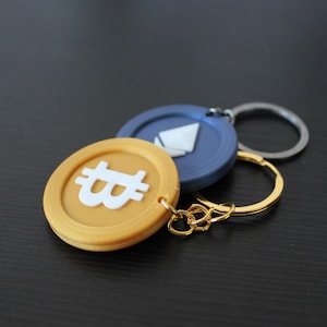 Può includere: Portachiavi dorati e blu con simboli di criptovaluta. Il portachiavi dorato ha un simbolo Bitcoin bianco e il portachiavi blu ha un simbolo Ethereum bianco.