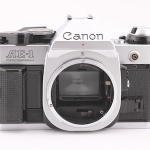 Può includere: Una fotocamera Canon AE-1 Program argento e nera. Il corpo macchina è argento con dettagli neri e presenta il logo Canon. L'attacco dell'obiettivo è aperto, rivelando i componenti interni. La fotocamera ha un design classico e vintage.