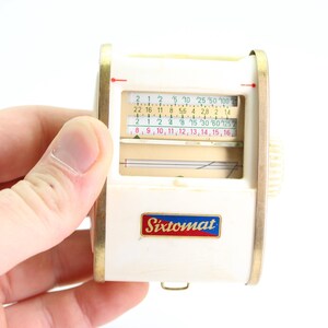 May include: A white and gold Sixtomat light meter with a sliding scale and a needle. The scale has numbers from 2 to 100 and 8 to 16.