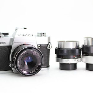 Può includere: Una macchina fotografica a pellicola da 35 mm di colore argento e nero con un obiettivo Topcon e due obiettivi aggiuntivi. La fotocamera ha un'etichetta Unirex sul davanti.