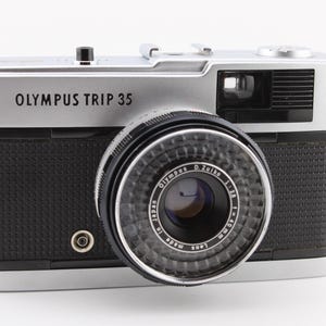 Op de afbeelding: Zilveren en zwarte Olympus Trip 35 camera met de tekst "OLYMPUS TRIP 35" op de voorkant. De camera heeft een zwarte, getextureerde grip en een lens met de tekst "Olympus D. Zuiko 1:2.8 f=40mm".