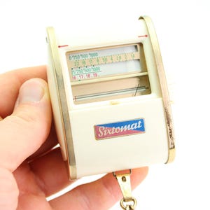 May include: A white and gold Sixtomat light meter with a black and white dial and a red and blue label. The light meter has a small chain attached to it.