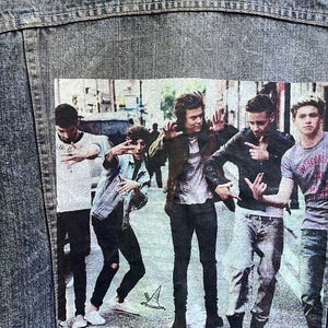 Op de afbeelding: Een spijkerjack met een bedrukte afbeelding van vijf mannen in een band. De afbeelding toont de mannen die op een straat in de stad staan, met gebouwen en auto's op de achtergrond.