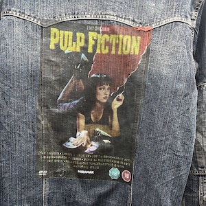 May include: A blue denim jacket with a Pulp Fiction movie poster patch sewn on the back. The patch features a woman lying down with a cigarette in her hand. The text on the patch reads "Pulp Fiction" and "Miramax".