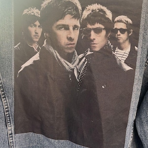 Może przedstawiać: Niebieska dżinsowa kurtka z czarno-białym naszywką zespołu na plecach. Naszywka przedstawia czterech mężczyzn z tekstem "BRITPOP XTREME" na metce nad nimi.