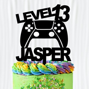 May include: A green cake with rainbow frosting and sprinkles. A black cake topper with a video game controller and the text "Level 13 Jasper" is on top of the cake.