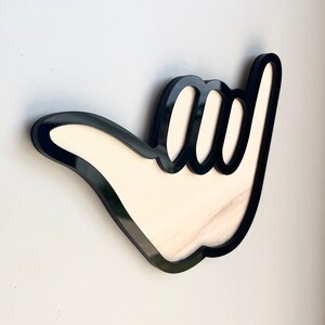 May include: Black and white wooden wall decor in the shape of a hand giving the shaka sign. The hand is outlined in black and the inside is white.
