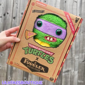 Op de afbeelding: Een Donatello-knuffel uit de Teenage Mutant Ninja Turtles x Fuggler-samenwerking, in een kartonnen doos. De knuffel heeft een groen gezicht, een paars masker en grote zwarte ogen. De doos bevat het Nickelodeon-logo, de tekst "Special Delivery" en een pizzapunt.