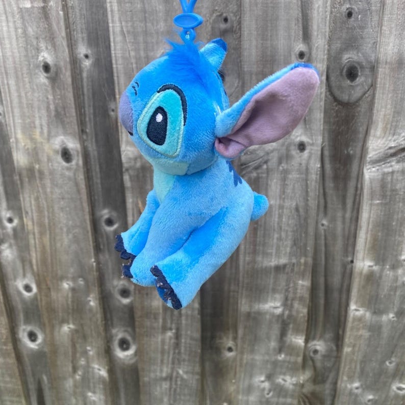 May include: A blue plush toy of the character Stitch, featuring large ears and a cheerful face. The toy is attached to a blue clip. The background is a wooden fence with a weathered appearance.
