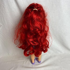 Puede incluir: Una mu&ntilde;eca con pelo largo y rojo vibrante peinado con una peque&ntilde;a goma de pelo de color claro. La mu&ntilde;eca lleva sandalias moradas. El pelo de la mu&ntilde;eca est&aacute; peinado con rizos sueltos.