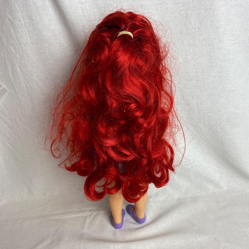 Puede incluir: Una mu&ntilde;eca con pelo largo y rojo vibrante peinado con una peque&ntilde;a goma de pelo de color claro. La mu&ntilde;eca lleva sandalias moradas. El pelo de la mu&ntilde;eca est&aacute; peinado con rizos sueltos.