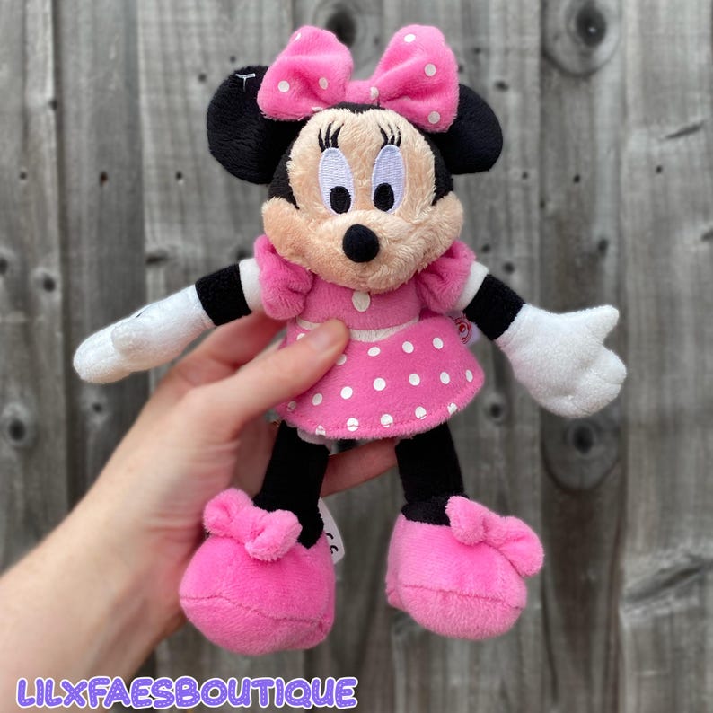 May include: A Minnie Mouse plush toy, featuring a pink dress with white polka dots, a matching bow, and pink shoes. The plush has black ears, a black nose, and white gloves. The text "LILXFAESBOUTIQUE" is visible at the bottom.