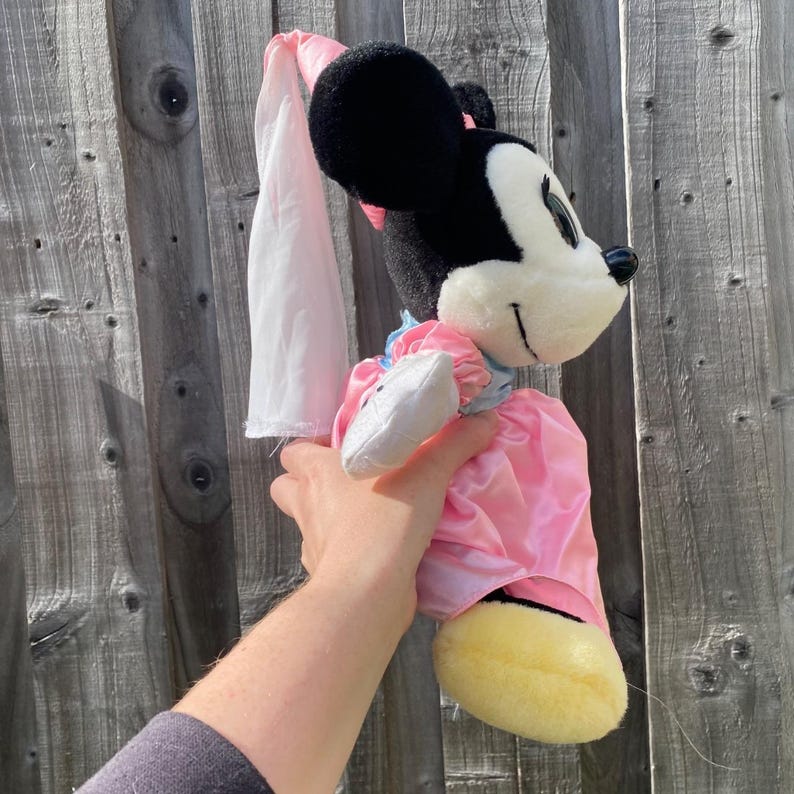 May include: A plush Minnie Mouse toy featuring a black head with large round ears, a white face, and a black nose. The character wears a shiny pink satin dress with light blue puffed sleeves and a long, flowing white veil attached to the head.