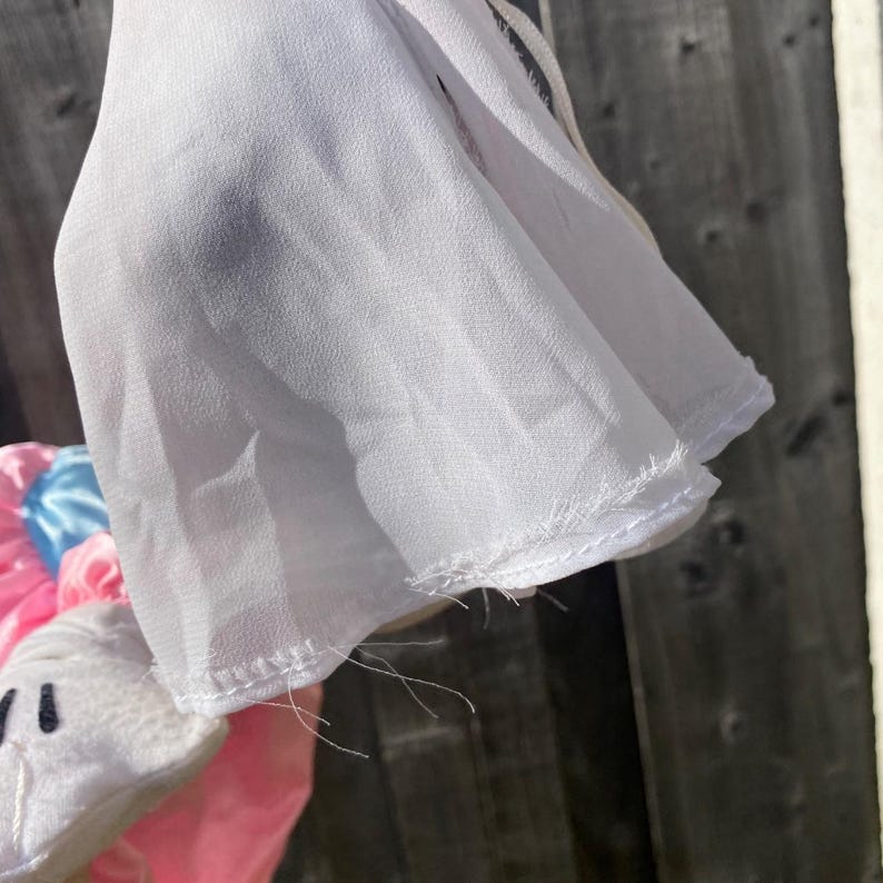 May include: A sheer white fabric sleeve with a stitched hem and loose threads, layered over a pink and blue satin garment. The white fabric has a translucent quality and a visible texture, showing the construction of the sleeve edge and seams.