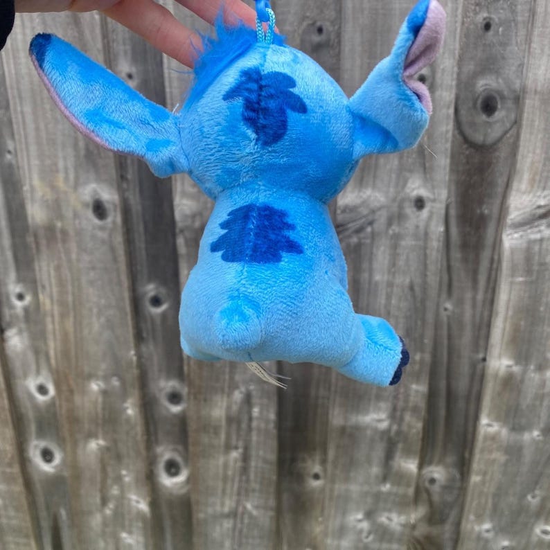 May include: A blue Stitch plush toy, featuring large ears, a tuft of hair, and dark blue markings on its back. The toy is suspended by a blue string. The toy is a popular character from Lilo & Stitch.