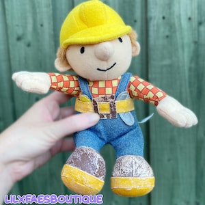 Peut inclure: Une peluche représentant un ouvrier du bâtiment portant un casque jaune vif, une chemise à manches longues à carreaux orange et jaune, et une salopette en jean bleu avec une boucle de ceinture marron. Les bottes marron ont un aspect usé.