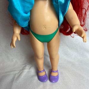 Puede incluir: Una mu&ntilde;eca con cabello rojo, que lleva una parte superior turquesa, una parte inferior verde y zapatos morados. La piel de la mu&ntilde;eca es de color bronceado claro. El cabello de la mu&ntilde;eca es largo y fluido.