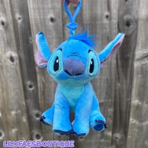 May include: A blue Stitch plush keychain with big black eyes, a purple nose, and large ears with pink inner ears. The keychain has a blue clip and the text "LILXFAESBOUTIQUE" at the bottom.