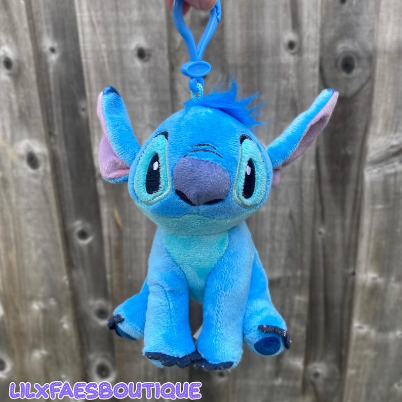 May include: A blue Stitch plush keychain with big black eyes, a purple nose, and large ears with pink inner ears. The keychain has a blue clip and the text "LILXFAESBOUTIQUE" at the bottom.